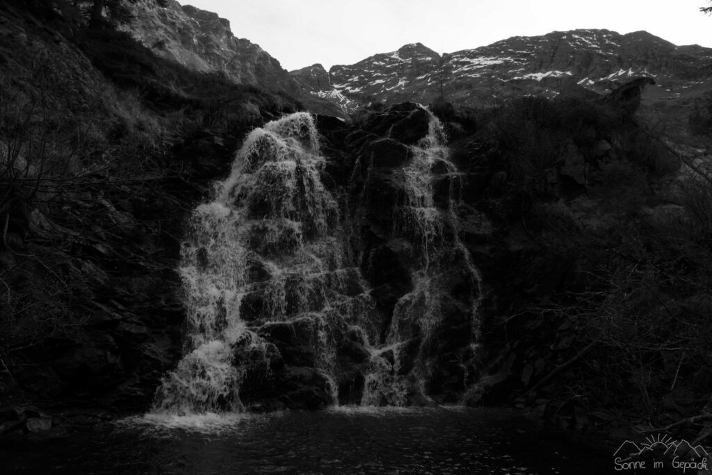 Schwarz-weiß Aufnahme von Wasserfall mit Bergen im Hintergrund.